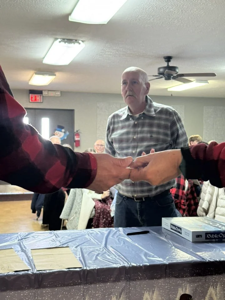 Two people shaking hands across a table with a Bible-themed board game on it, a man with a gray plaid shirt standing in the background, in a room decorated for a gathering with people seated at tables.