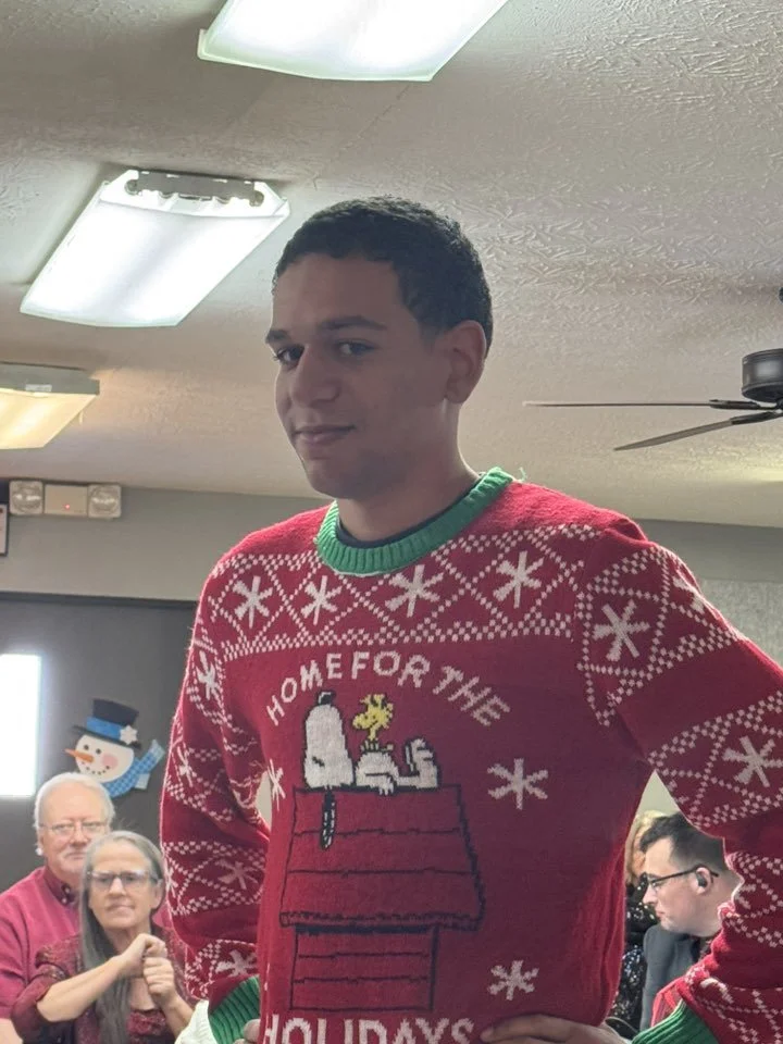 A young man wearing a Christmas sweater with a graphic of Snoopy on his doghouse from Peanuts and the phrase "Home for the Holidays". In the background, several seated people are visible, including an older man with glasses and a woman with long hair