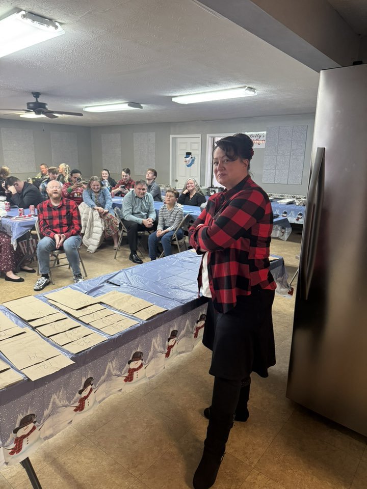 A woman in a red and black plaid shirt standing next to a table with wrapped items, facing an audience in a room decorated with snowmen-themed holiday decor.