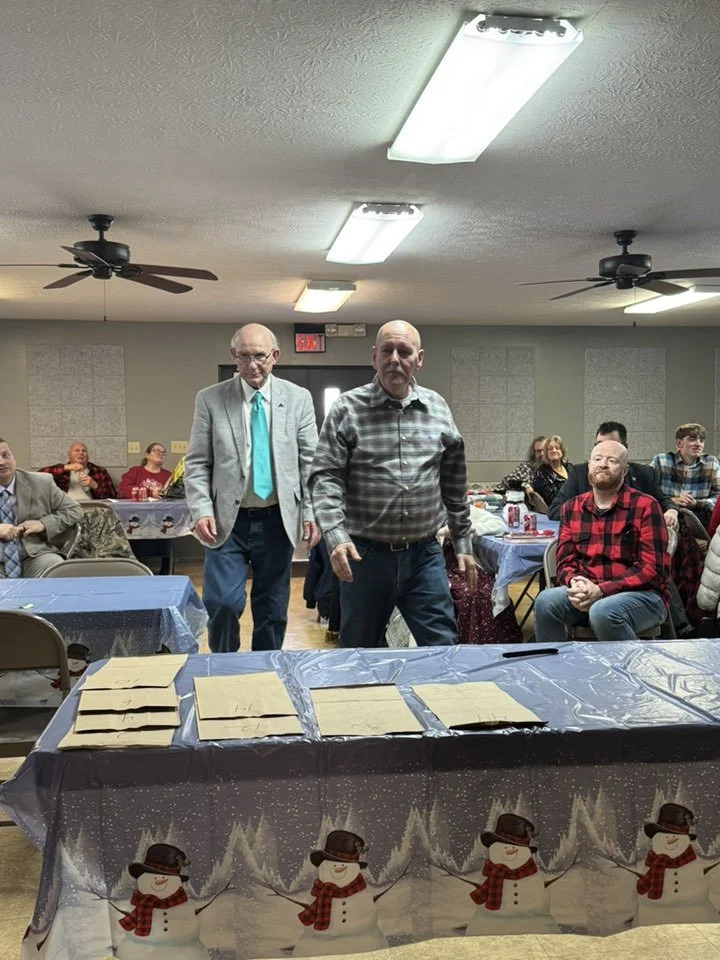People gathered in a decorated community hall for a Christmas event. Two men stand in the center, one in a gray blazer and blue tie, the other in a plaid shirt. Several people are seated at tables in the background, some wearing festive clothing. The
