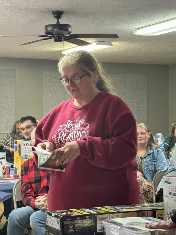 Woman with glasses, wearing a red sweatshirt with white text, reading a booklet in a room with people sitting at tables.