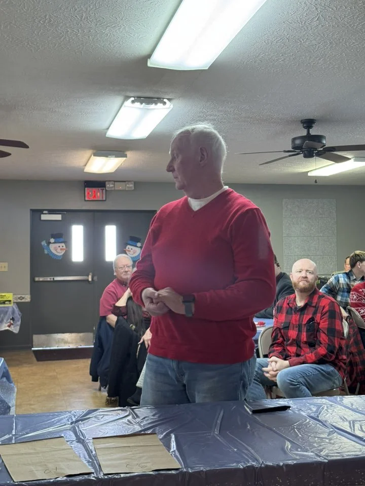 An elderly man in a red sweater standing at a table during a social gathering in a decorated room, with people seated at tables around him.