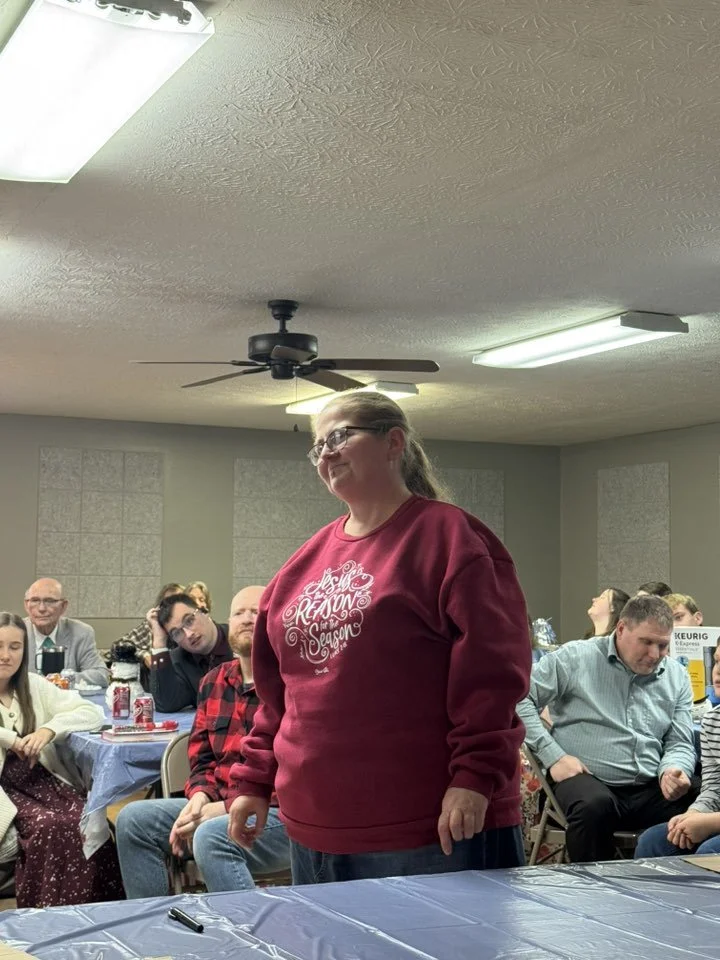 A woman with glasses wearing a red sweatshirt standing in a room with a group of people sitting at tables, some looking at her. The sweatshirt has holiday-themed text. The room has a white ceiling with ceiling fans and fluorescent lights.