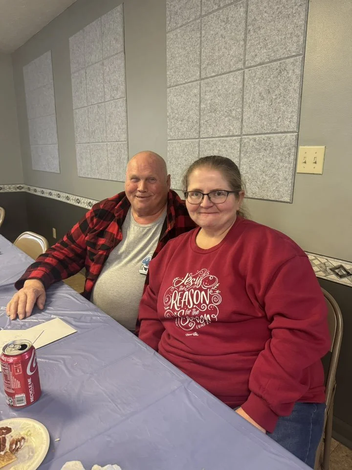 A man and woman sitting at a table with a blue tablecloth, smiling at the camera. The man is wearing a gray shirt with a red and black plaid shirt over it, and the woman is wearing glasses and a red sweatshirt with white lettering. The background sho