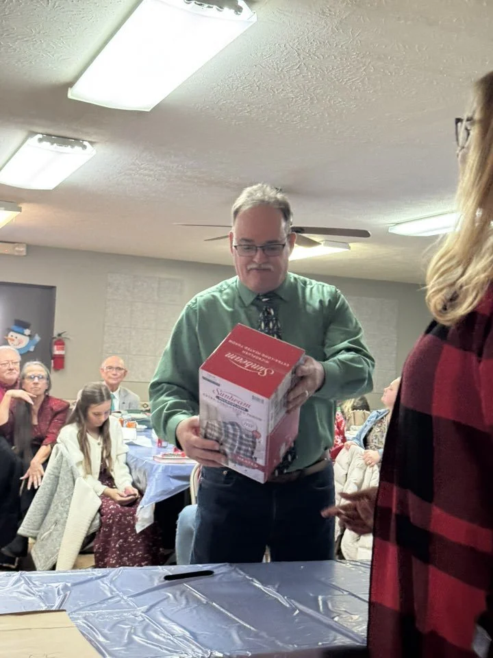 Man in green shirt and glasses opening a box at a gathering with several seated people observing.