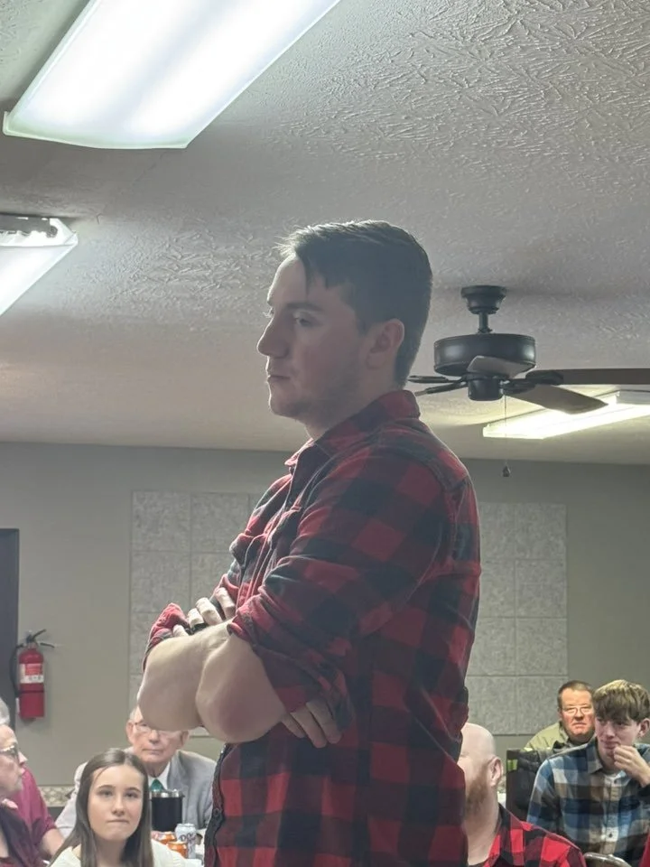 A young man with dark hair and light skin standing with arms crossed, wearing a red and black plaid shirt, in a room with a textured ceiling, fluorescent lights, a ceiling fan, and a group of people sitting at tables.