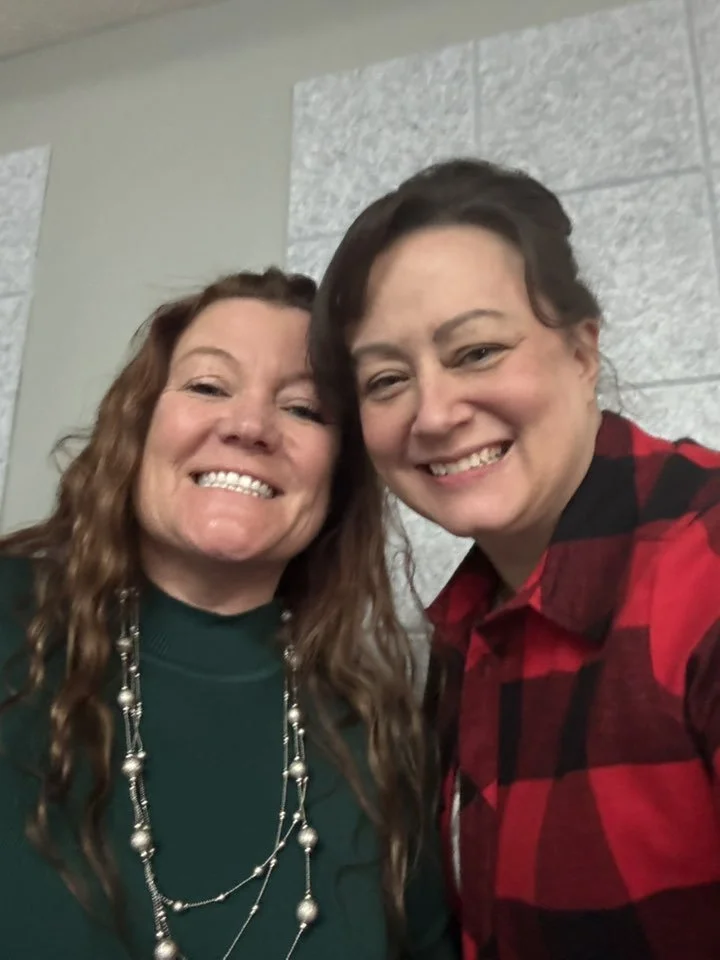 Two women smiling and posing for a selfie indoors, against a wall with textured white panels.