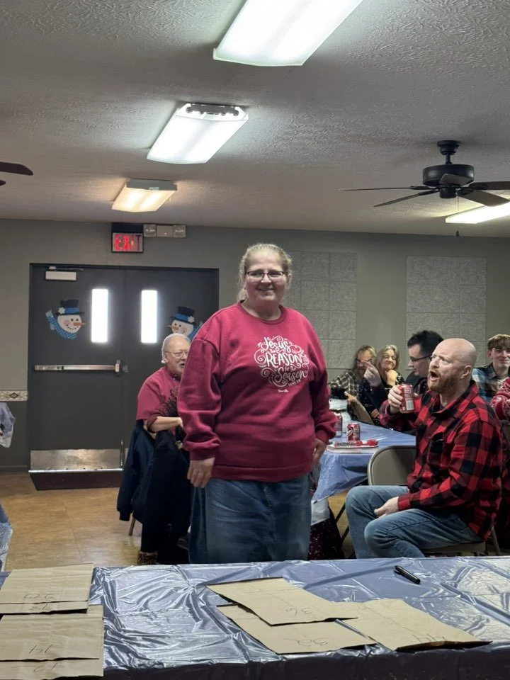 A group of people, mostly middle-aged adults, sitting at tables decorated with blue tablecloths and Christmas-themed decorations, gathered in a room with Christmas snowman door decals. A woman in a red sweater stands in the foreground, smiling, with 
