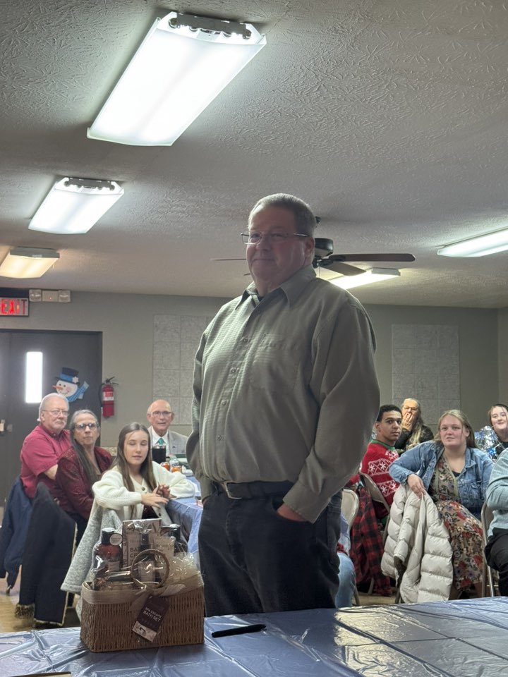 A man standing in a room full of seated people, with a table in front of him and ceiling lights above.