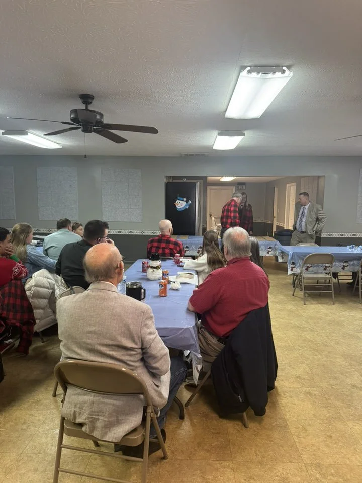 A group of people seated at tables with blue tablecloths in a room, listening to two men speaking at the front near a door decorated with a snowman. The room has ceiling fans and fluorescent lights, with some decorations on the walls.