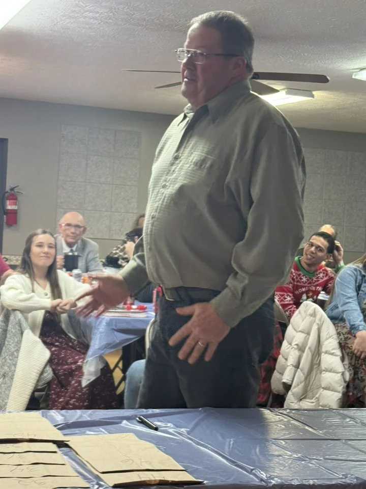 A man wearing glasses and a gray button-up shirt shakes hands with someone in a crowded indoor gathering, with people sitting at tables in the background.
