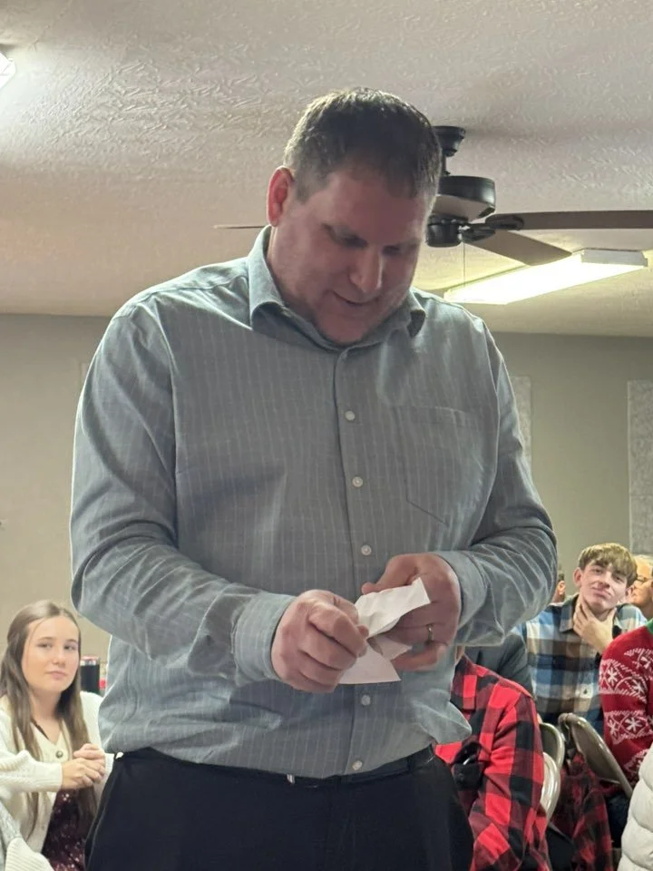 A man in a gray shirt standing and holding a tissue, smiling. Several people are seated behind him, including a girl in a white sweater and other young individuals, in an indoor setting with ceiling fan and lights.