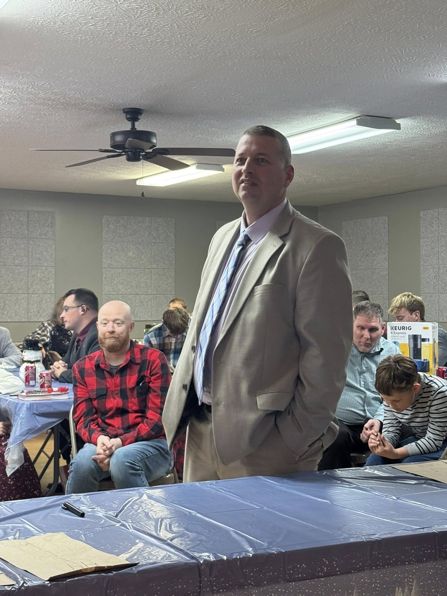 Man in beige suit standing in a room with other seated people at tables, some with soda cans, in a room with a low ceiling and fluorescent lighting.
