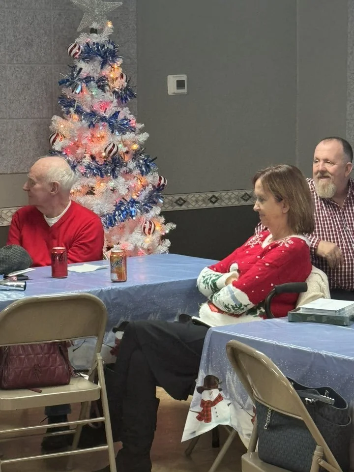 People sitting at a table near a decorated Christmas tree, with some wearing red and festive sweaters, in an indoor setting.