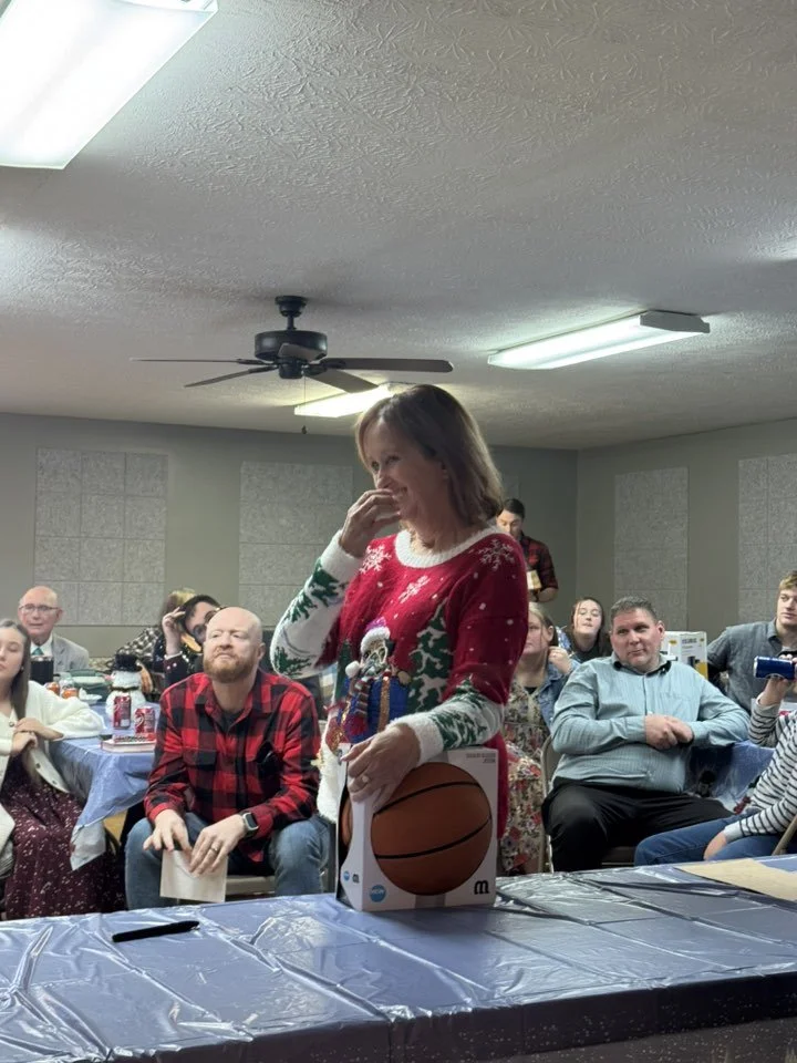 Woman in a Christmas sweater standing in front of a table with a basketball gift box, smiling and covering her mouth in a room with people seated and watching.
