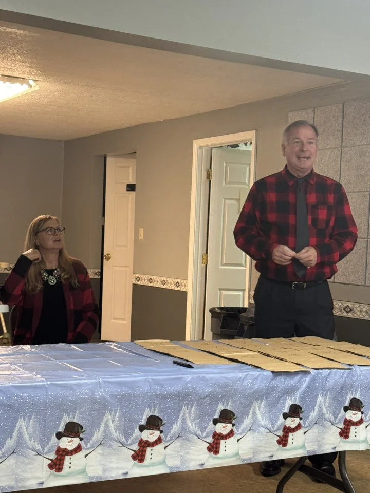 A man in a red and black plaid shirt and tie stands at a table covered with paper bags, smiling while speaking to an audience. A woman in a matching plaid shirt and glasses sits at the table, listening.