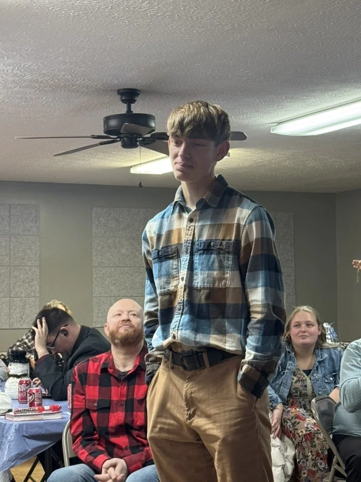 Young man standing in a room with several seated people, some looking at him with smiles, under a ceiling fan and fluorescent lights.