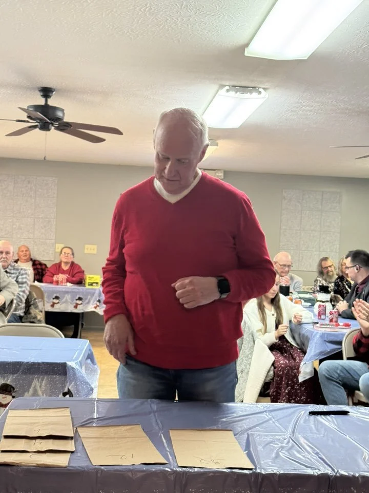 A man wearing a red sweater and watch standing at a table with brown paper bags, with people seated at decorated tables in the background celebrating in a community hall.
