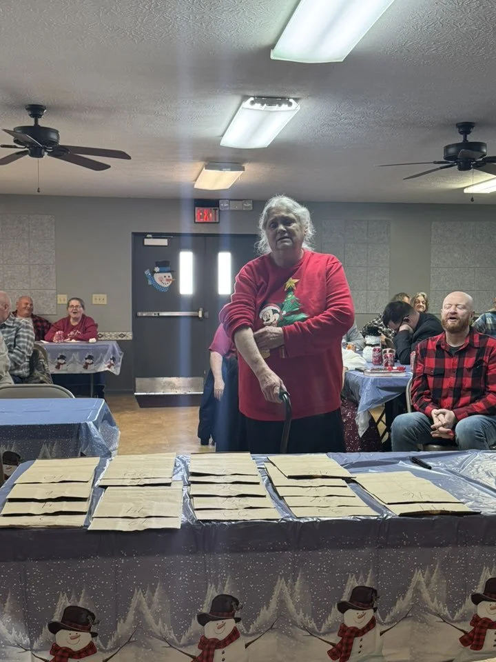 A woman in a red Christmas sweater standing behind a table with wrapped presents, at a holiday gathering with seated guests in a decorated room.