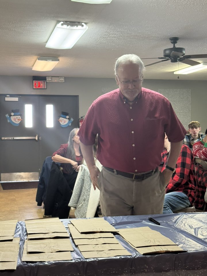 An older man with white hair and glasses, wearing a red button-up shirt and khaki pants, stands at a table covered with paper bags. In the background, there are holiday decorations on the door, including snowmen, and people sitting at tables in a roo