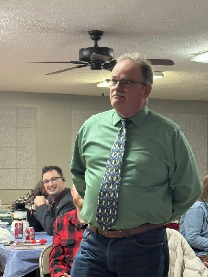 A man with glasses and gray hair standing with his arms behind his back, wearing a green shirt and patterned tie, in a room with a ceiling fan and several people sitting at a table, smiling and engaging.