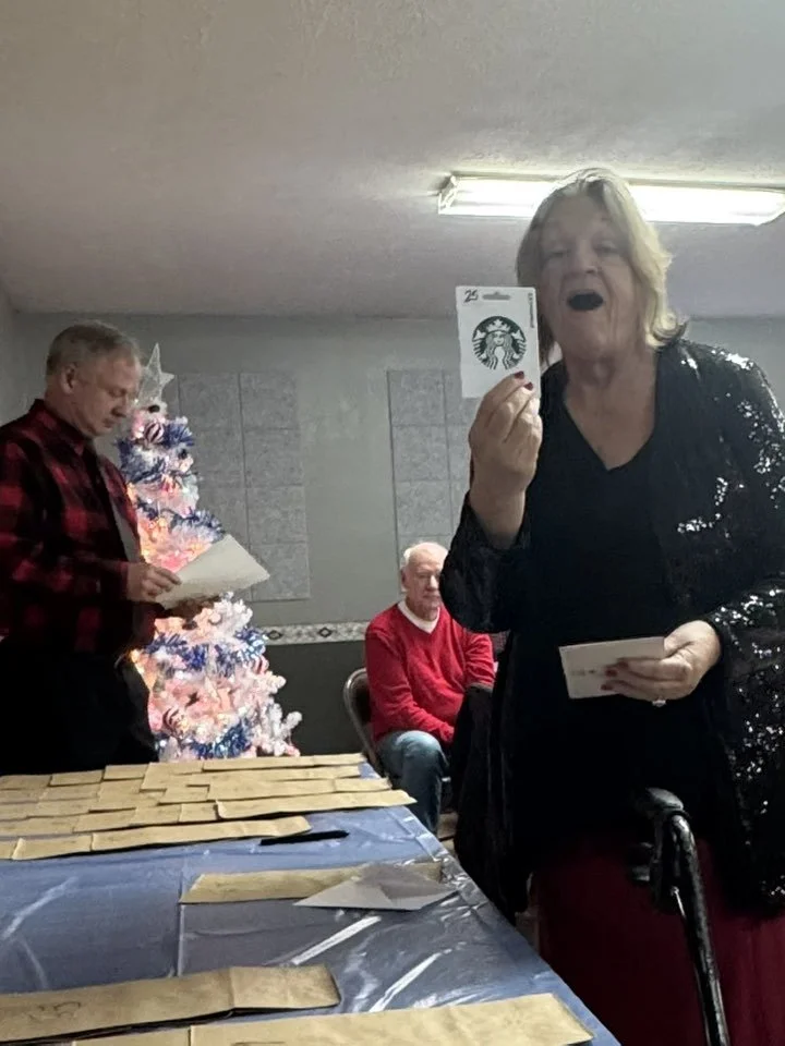 Elderly woman at a gathering holding a Starbucks gift card and smiling, with a Christmas tree decorated with pink, white, and blue ornaments in the background, and a man in a red sweater sitting nearby.