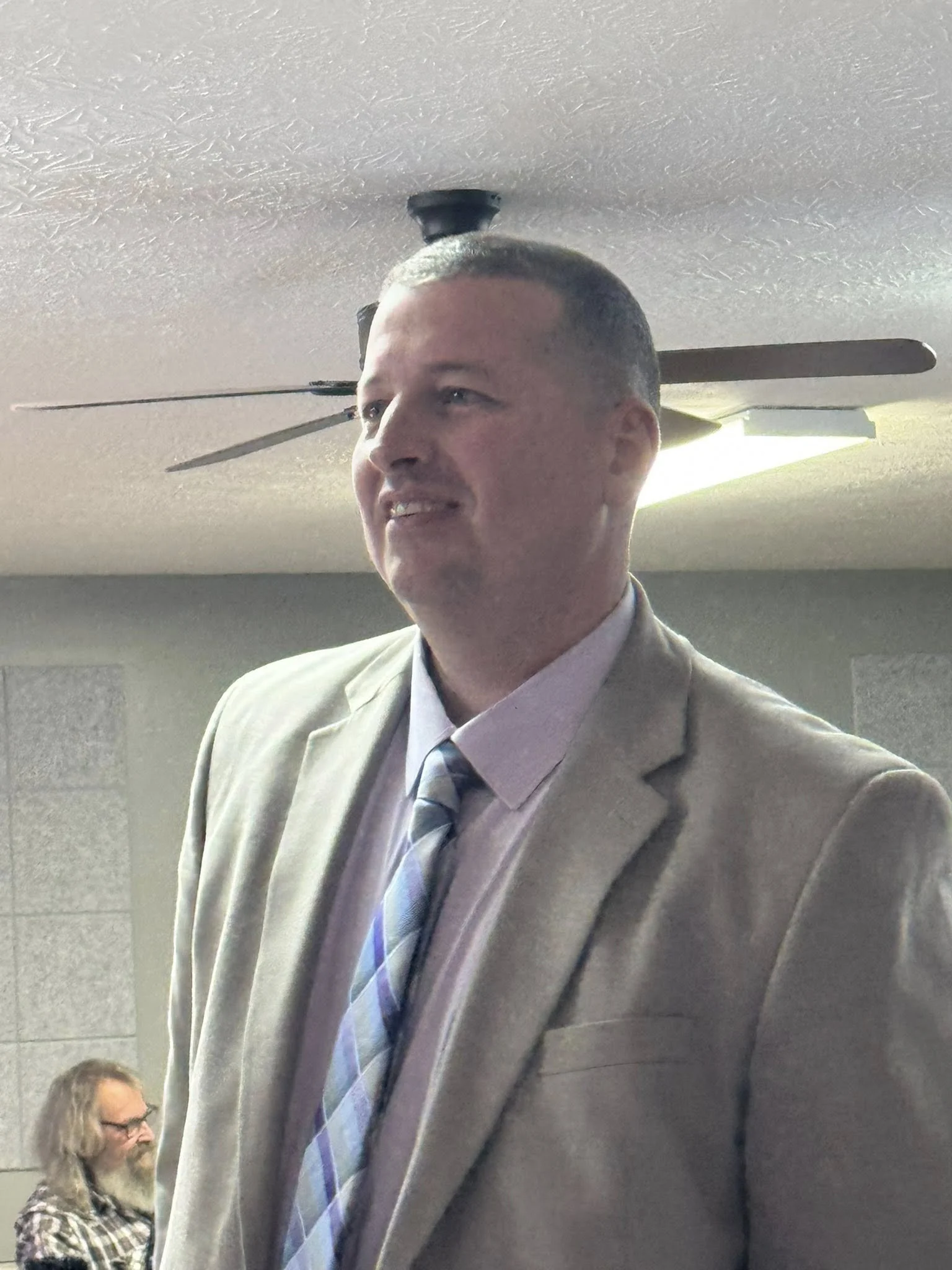A man in a beige suit and a striped tie standing indoors with a ceiling fan above and a person with long hair and glasses sitting in the background.