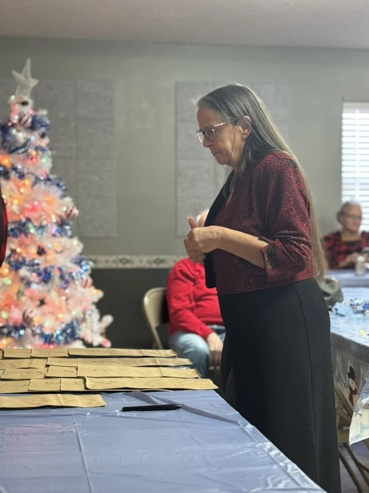 A woman with long gray hair, glasses, and a red and black dress or jacket stands at a table covered with brown paper gift bags during a holiday gathering. A decorated pink Christmas tree with ornaments and lights is in the background.