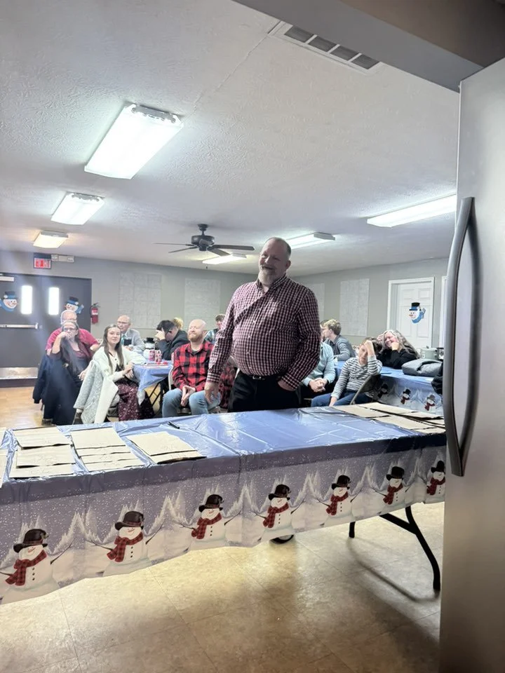 A group of people sitting at tables in a decorated room, with a man standing in front of a table covered with a snowman-themed tablecloth, inside a room with ceiling lights, a ceiling fan, and holiday decorations.