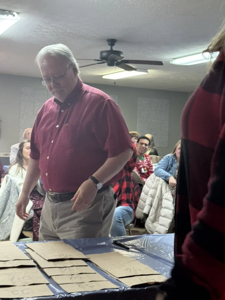 An indoor event with multiple people seated around tables. A man in a red shirt and beige pants is standing and looking down at a table with labeled papers. The room has gray walls, acoustic panels, a ceiling fan, and fluorescent lights.