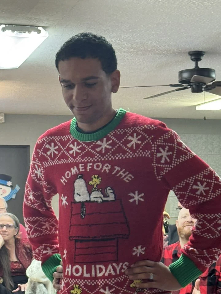 A man wearing a red Christmas sweater with Snoopy on top of his doghouse and the words 'Home for the Holidays'. He has dark hair and is smiling with his hands on his hips. Several people are in the background at what appears to be a holiday party.