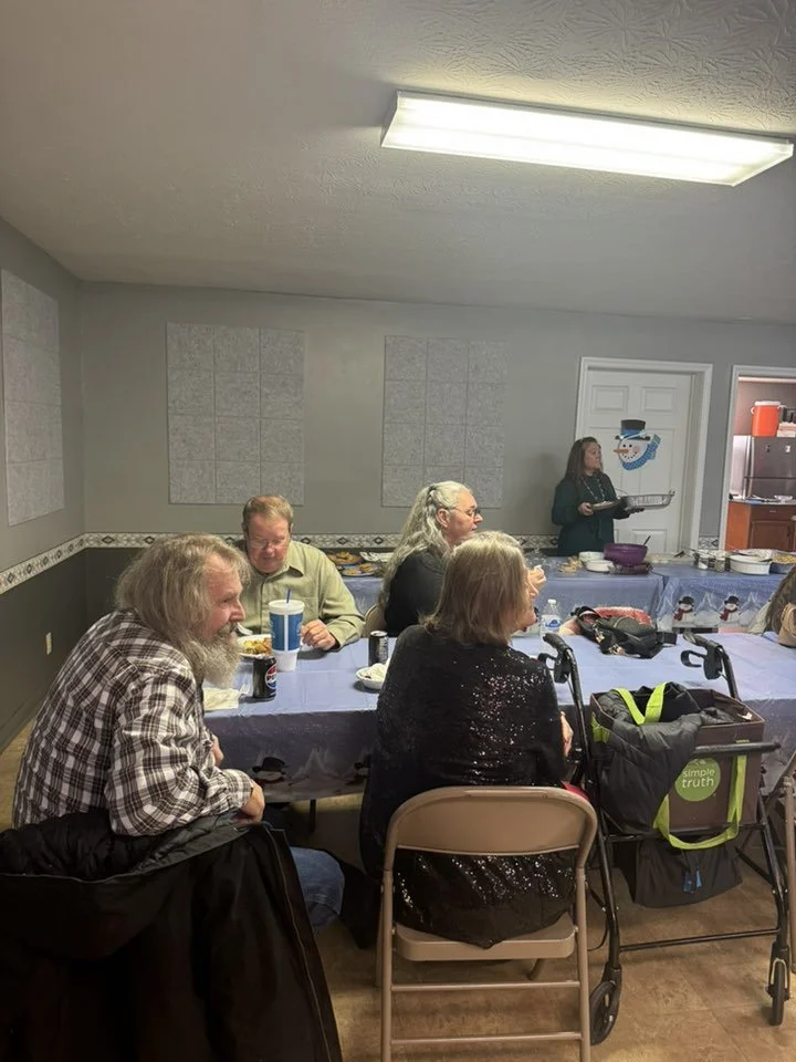 People gathered around a dining table with food, some using wheelchairs. A woman is serving food in the background, and the room has gray walls decorated for a holiday, possibly Christmas.