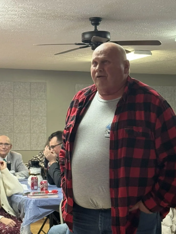 Bald man wearing a red and black plaid shirt standing indoors at a gathering, with other people sitting at a table behind him.