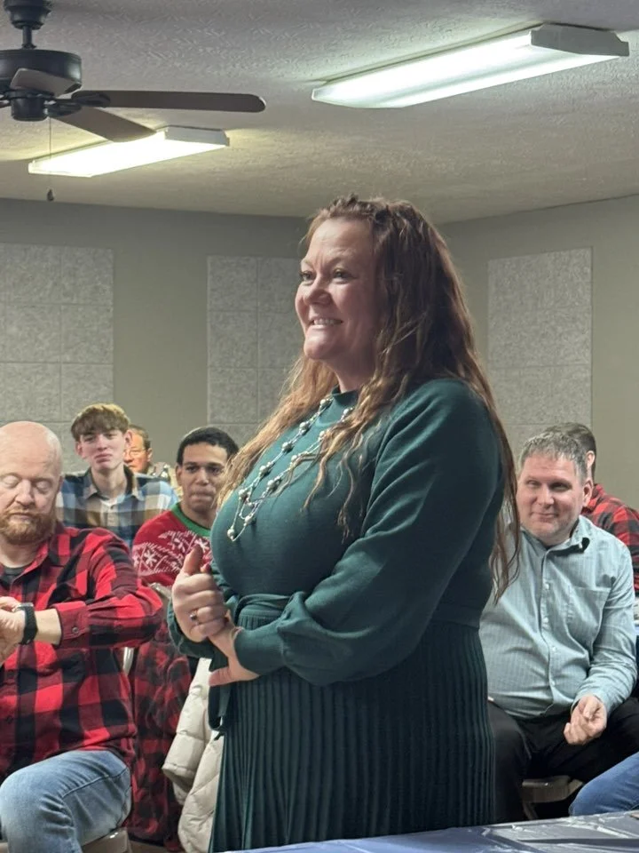 A woman in a green dress stands in front of an audience, smiling warmly. The audience includes several men and young people, some wearing plaid and festive sweaters, seated in a room with fluorescent lighting and a ceiling fan.