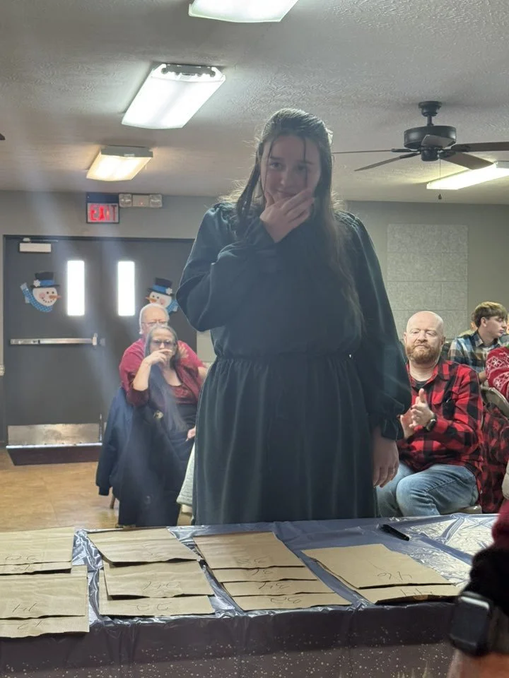A young woman standing in front of a table covered with brown paper bags, covering her mouth with her hand, in a room with holiday decorations and people sitting around, some clapping.