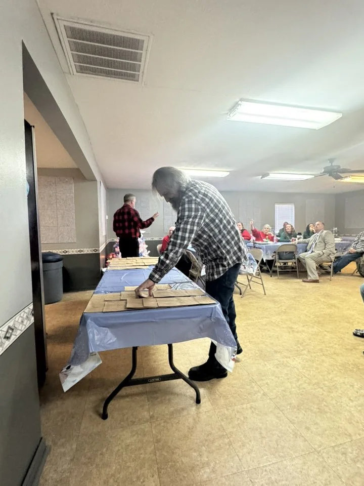 A man in checkered shirt standing over a table with many individually wrapped pieces of food, possibly cookies or crackers. There are several people seated at tables in the background in a large room with fluorescent lighting.
