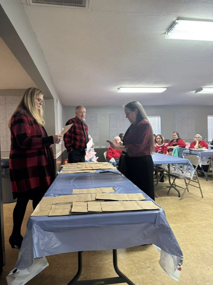 People gather around a table with brown paper bags, preparing for what appears to be a gift exchange or distribution event in a room decorated for a holiday celebration.