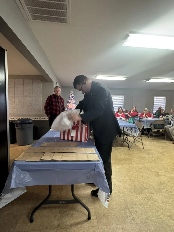 A man in a black suit opening holiday gift bags at a table, with a woman in a red and black plaid shirt standing behind him in a decorated indoor space. Several people sit at tables in the background.