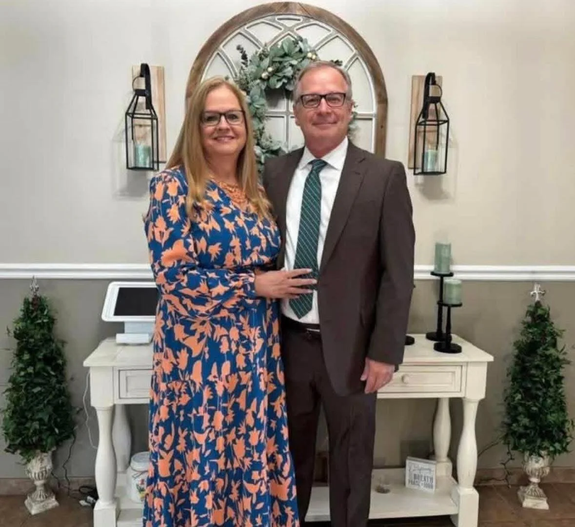 A man and a woman standing together indoors, smiling, in front of a decorative wall with a wreath and lanterns, with small trees on either side of a white table.