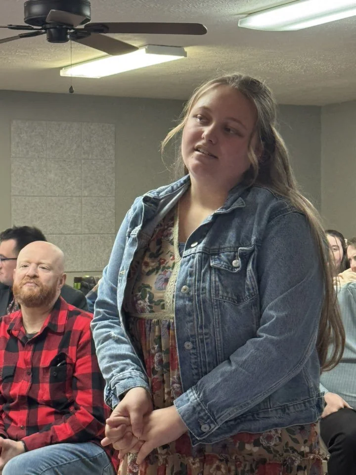 A young woman with long brown hair, wearing a floral dress and denim jacket, standing in a room with seated people, some paying attention, others not.