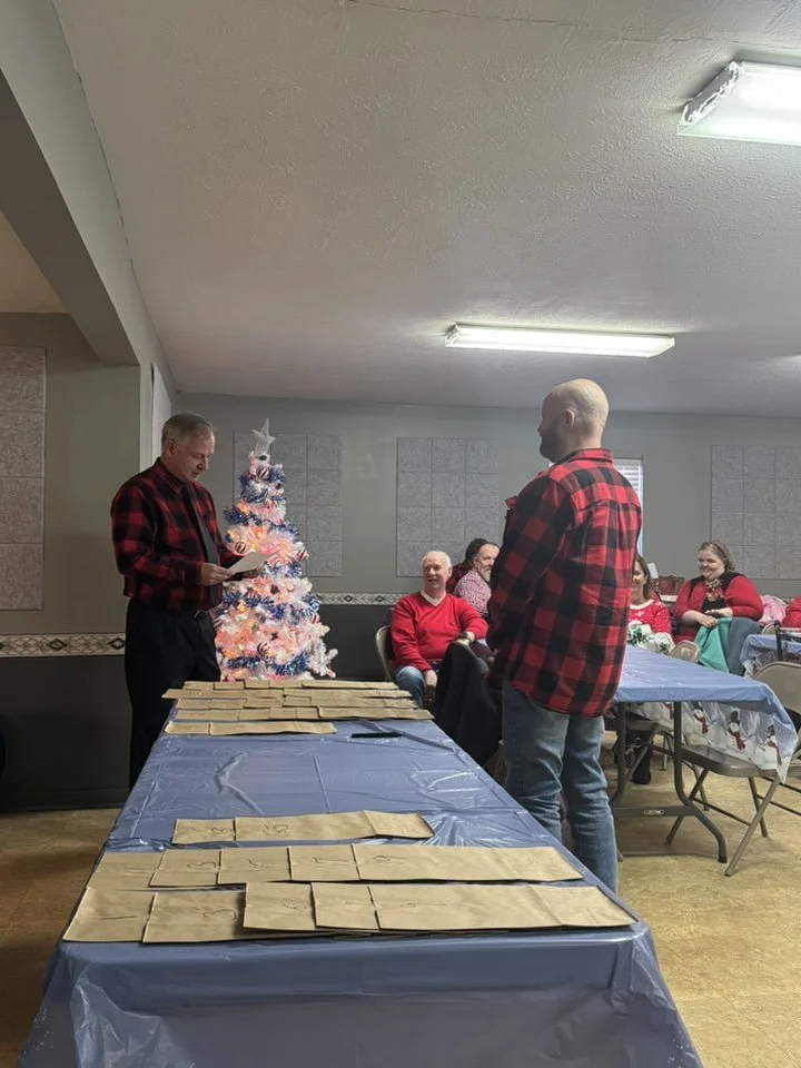 Man in red and black plaid shirt reading a paper, speaking to another man in similar plaid shirt, with a decorated Christmas tree in the background, in a decorated room with several seated people smiling.