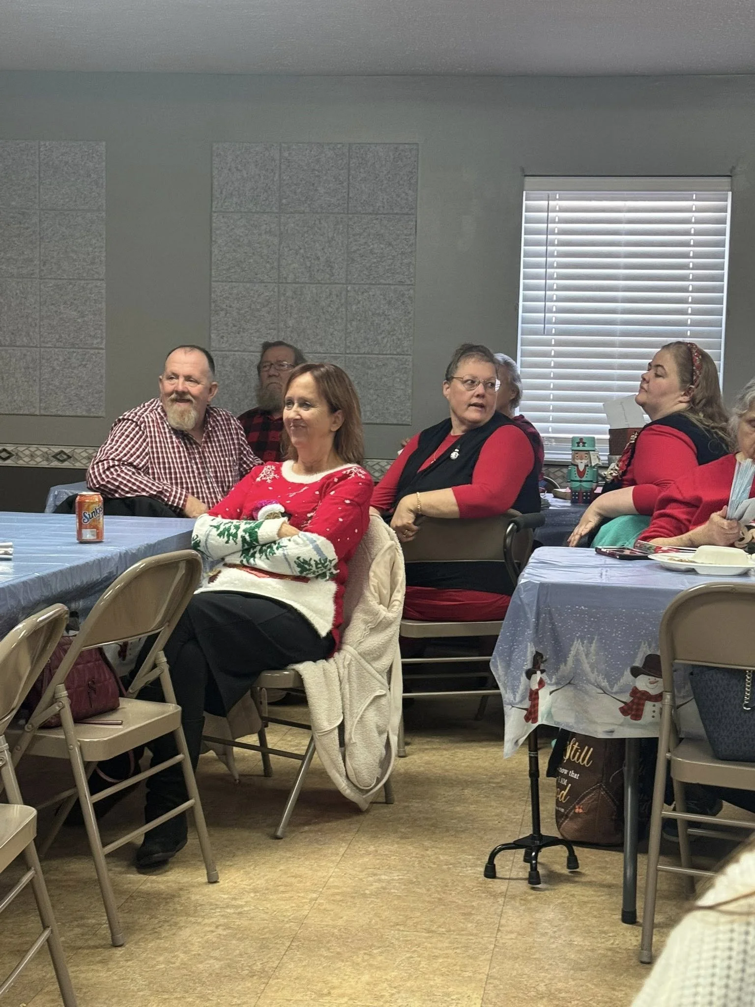 People sitting at tables during a holiday gathering, with Christmas decorations and festive clothing, in a room with gray walls and a window with blinds.