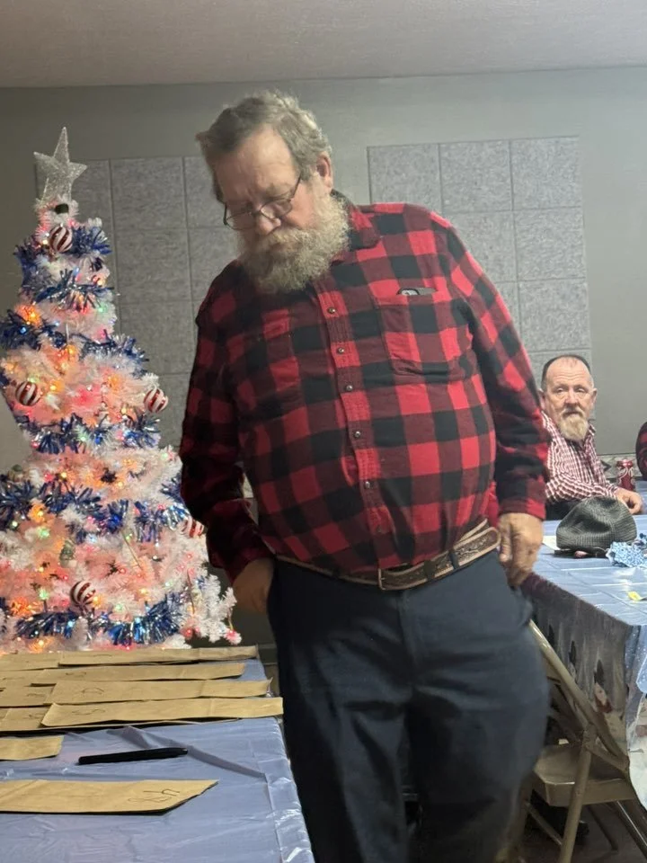 A man with glasses and a beard in a red and black plaid shirt standing in front of a decorated Christmas tree at a holiday gathering.