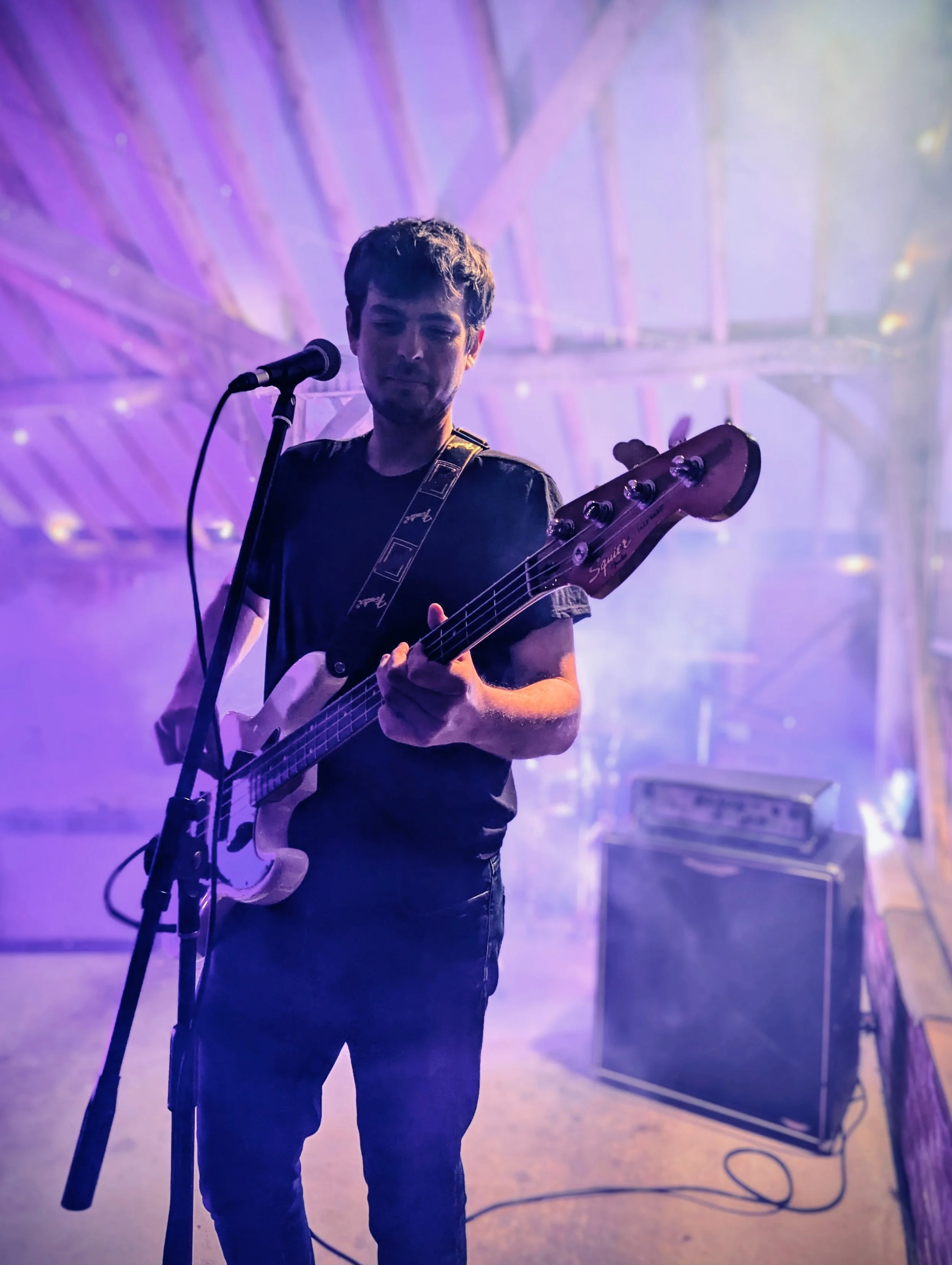 A young man playing an electric bass guitar on stage with purple and blue lights, smoke, and an amplifier in the background.
