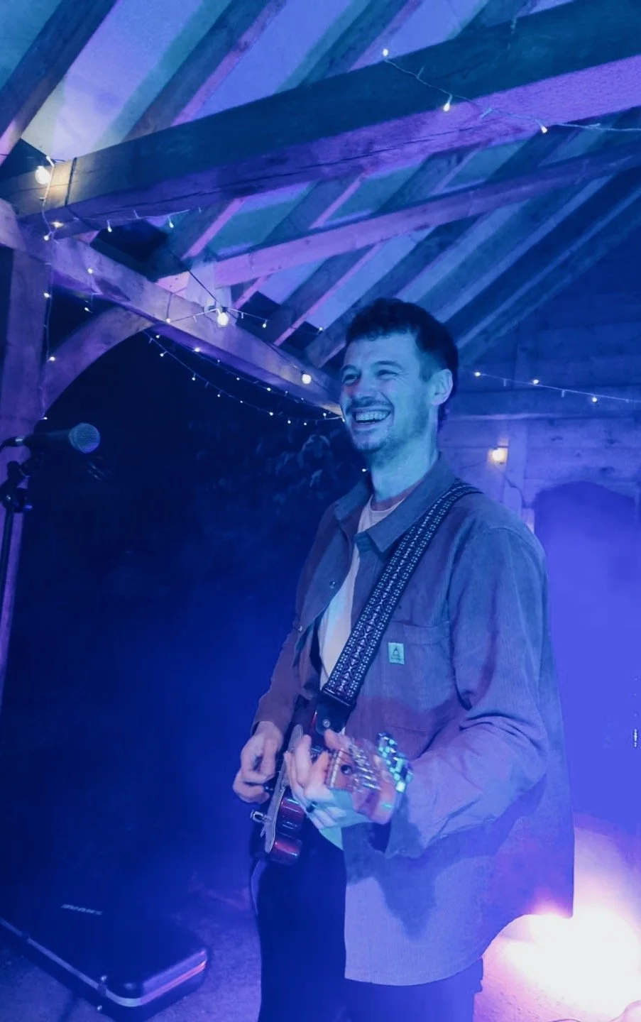 A young man with curly dark hair and a beard, smiling and playing an electric guitar on stage with purple and blue lighting, in a rustic wooden venue decorated with string lights.