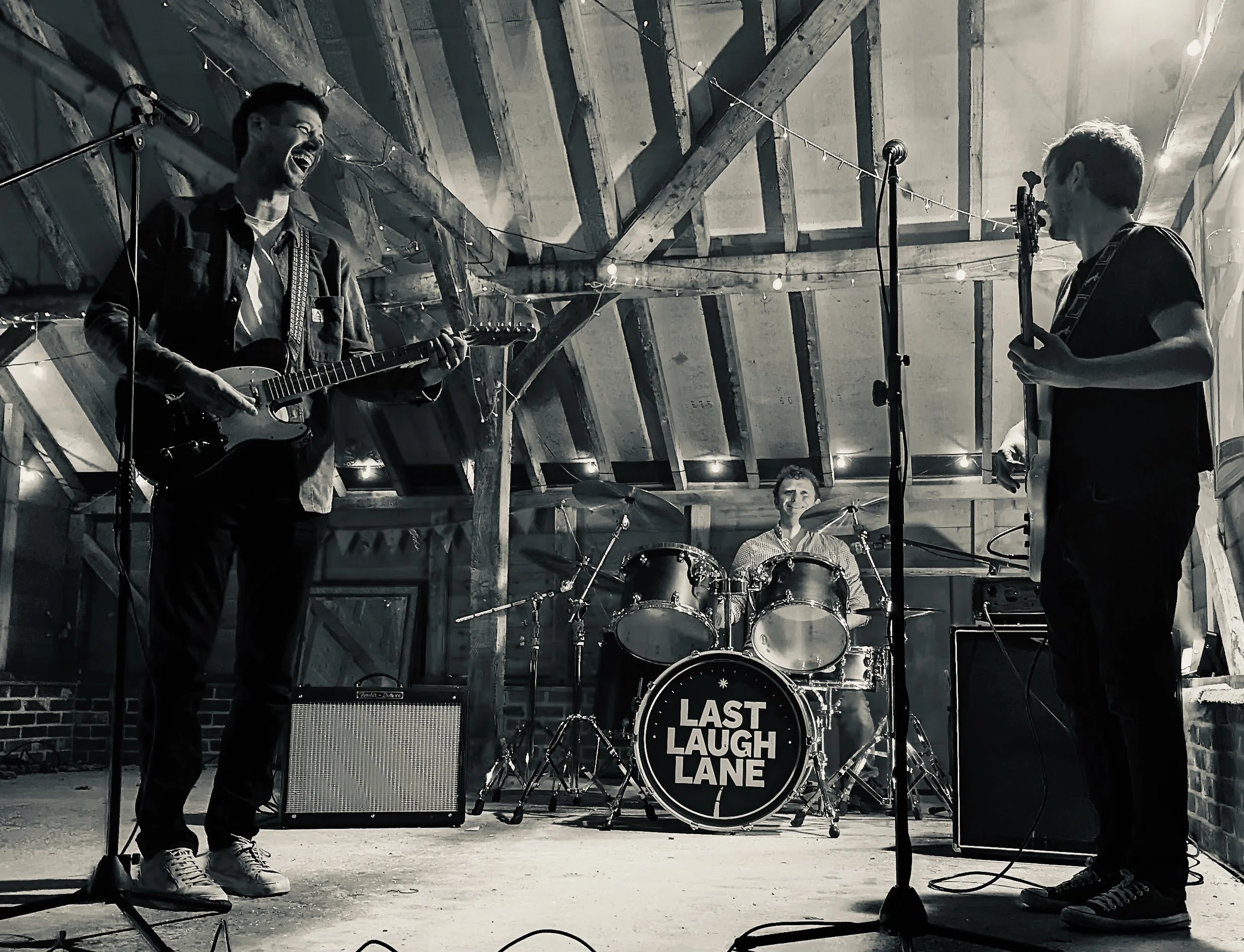Three band members performing on stage in a rustic venue with wooden beams, two guitarists in front and a drummer in the background, with 'Last Laugh Lane' on the drum kit.