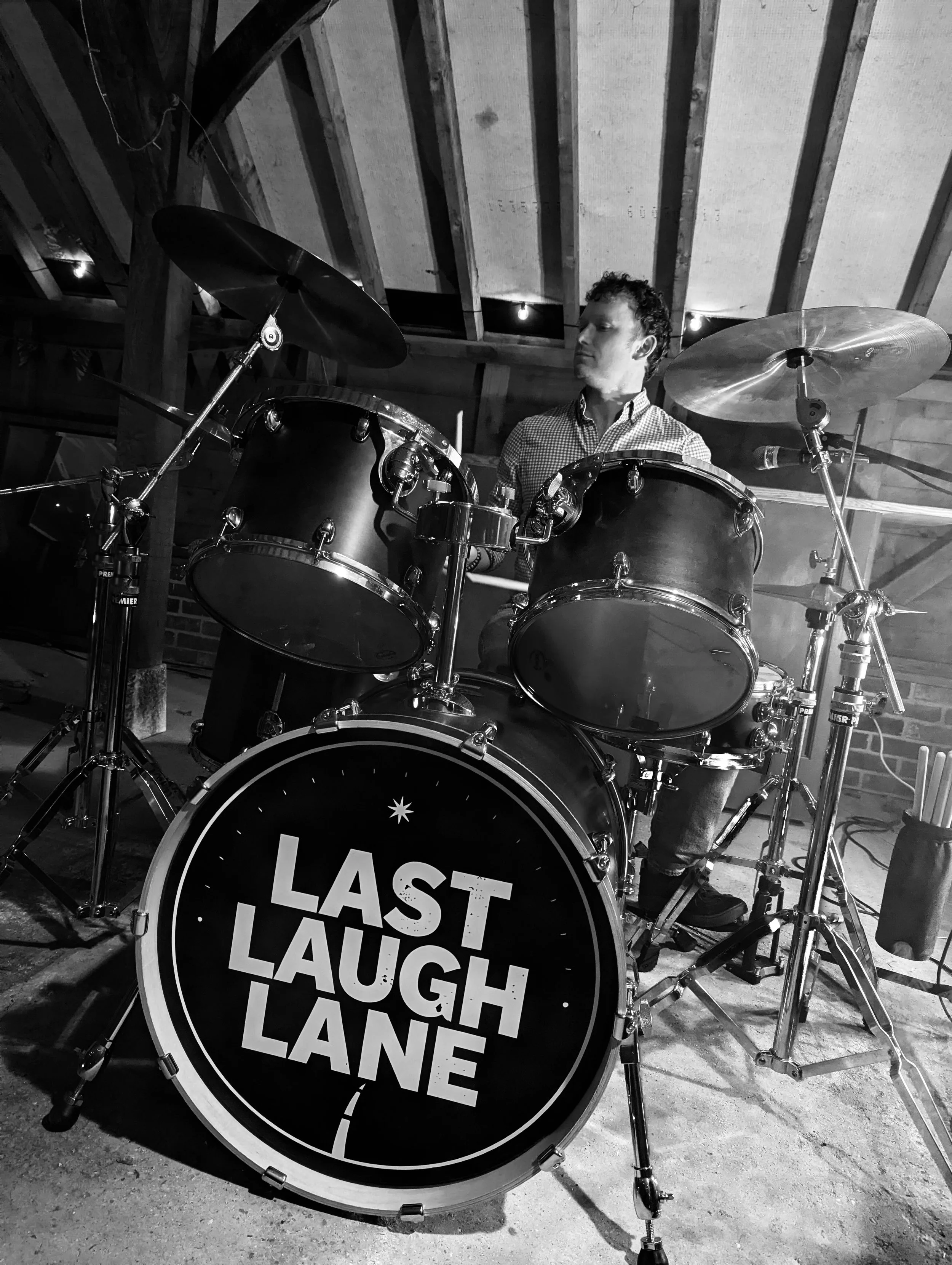 A man sitting at a drum set in a rustic wooden room with a pitched roof. The bass drum has the words 'LAST LAUGH LANE' written on it. The image is in black and white.