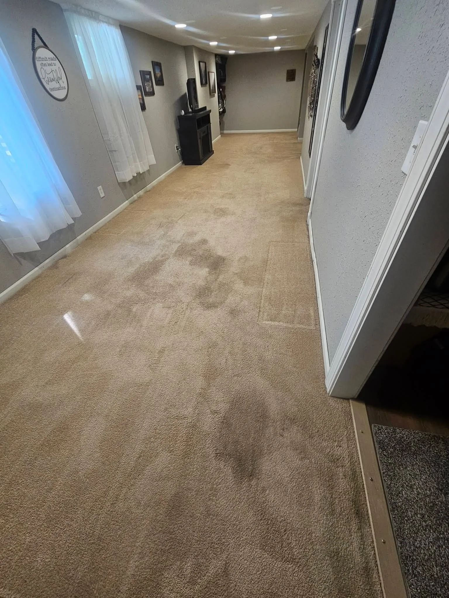 Empty hallway with beige carpet, white walls, and a black fireplace. There are framed pictures on the walls, white curtains on the window, and ceiling lights. Part of a door and a wall-mounted mirror are visible.