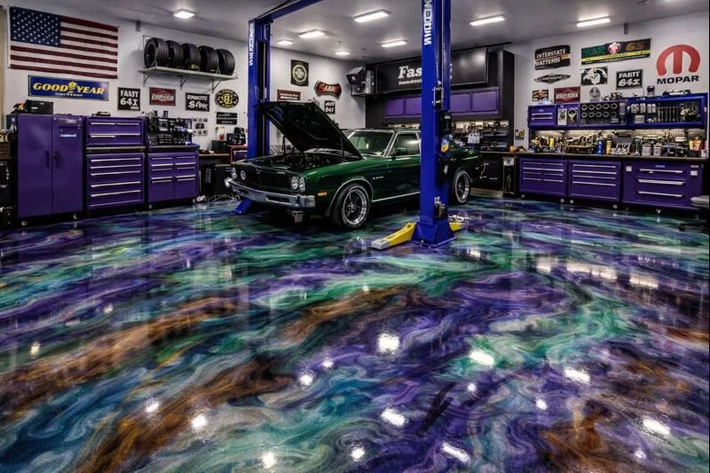 A green classic car with its hood open in a brightly lit auto repair garage with purple tool cabinets, brand signs, and a colorful, swirling epoxy floor.