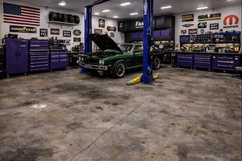 A green vintage muscle car with its hood open inside a well-equipped garage or workshop, featuring purple tool cabinets, automotive signage, and a vehicle lift.